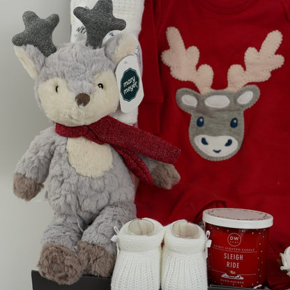 Christmas baby hamper, organic cotton red baby sleepsuit with applique reindeer, reindeer soft toy in grey and cream with a red scarft and glittery antlers, white cotton cellular baby blanket, white knit baby booties, red Christmas candle