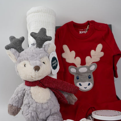 Christmas baby hamper, organic cotton red baby sleepsuit with applique reindeer, reindeer soft toy in grey and cream with a red scarft and glittery antlers, white cotton cellular baby blanket, white knit baby booties, red Christmas candle