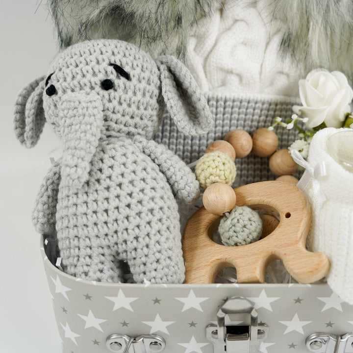 Grey gift suitcase with white stars, cream baby hat with grey trim and grey fluffy double pom poms , grey amigurumi elephant and wooden elephant teether with wooden and crocheted beads , white baby booties