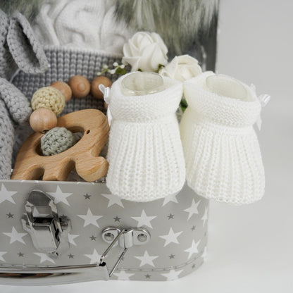 Grey gift suitcase with white stars, cream baby hat with grey trim and grey fluffy double pom poms , grey amigurumi elephant and wooden elephant teether with wooden and crocheted beads , white baby booties