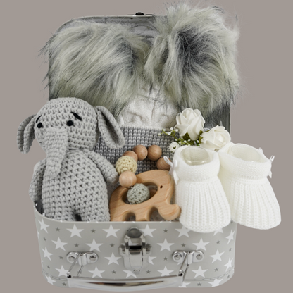 Grey gift suitcase with white stars, cream baby hat with grey trim and grey fluffy double pom poms , grey amigurumi elephant and wooden elephant teether with wooden and crocheted beads , white baby booties 