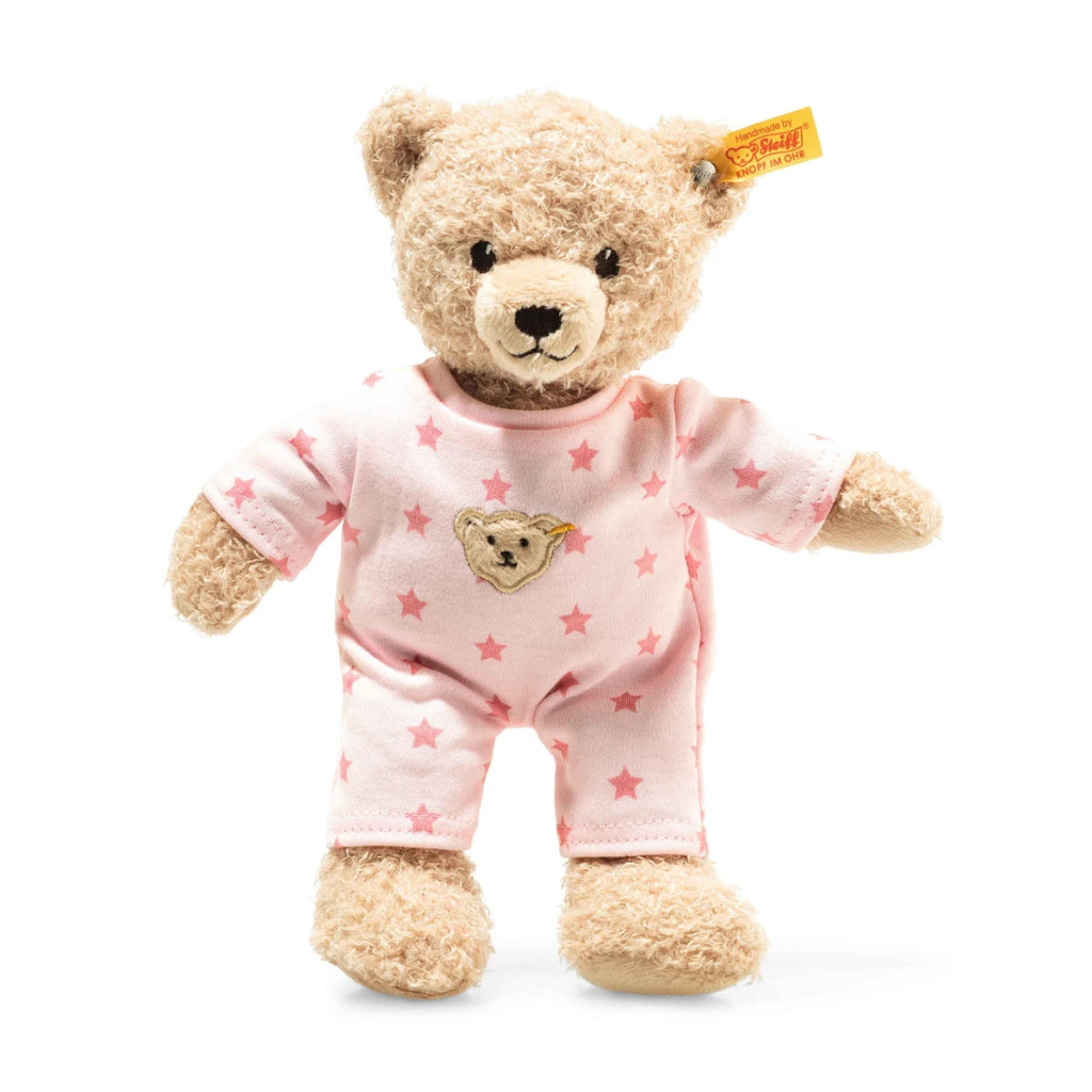 A plush teddy bear toy wearing light pink pajamas with darker pink stars and a bear face patch on the chest, photographed on a clean white background. A yellow Steiff brand tag is visible on the ear.