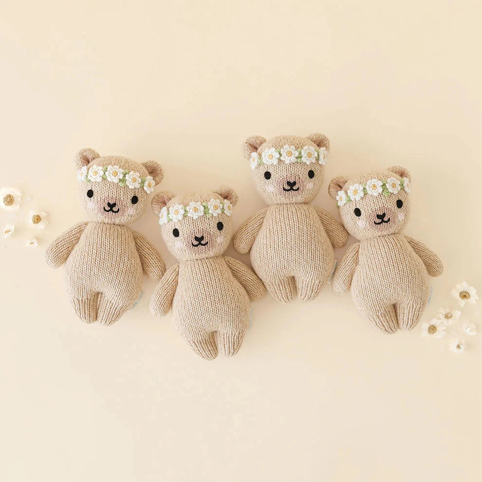 Four knitted teddy bears in different sizes, arranged in a row on a light beige background. Each bear wears a small white daisy flower crown.