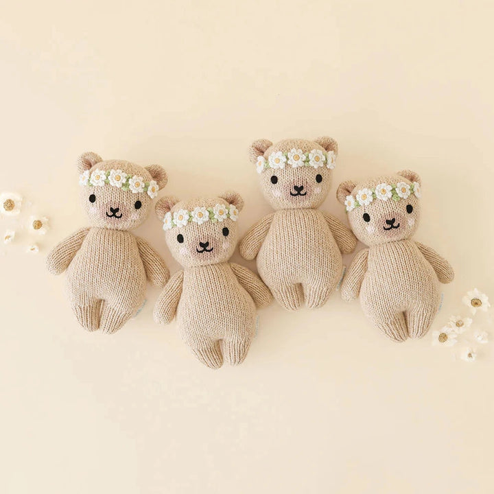 Four knitted teddy bears in different sizes, arranged in a row on a light beige background. Each bear wears a small white daisy flower crown.