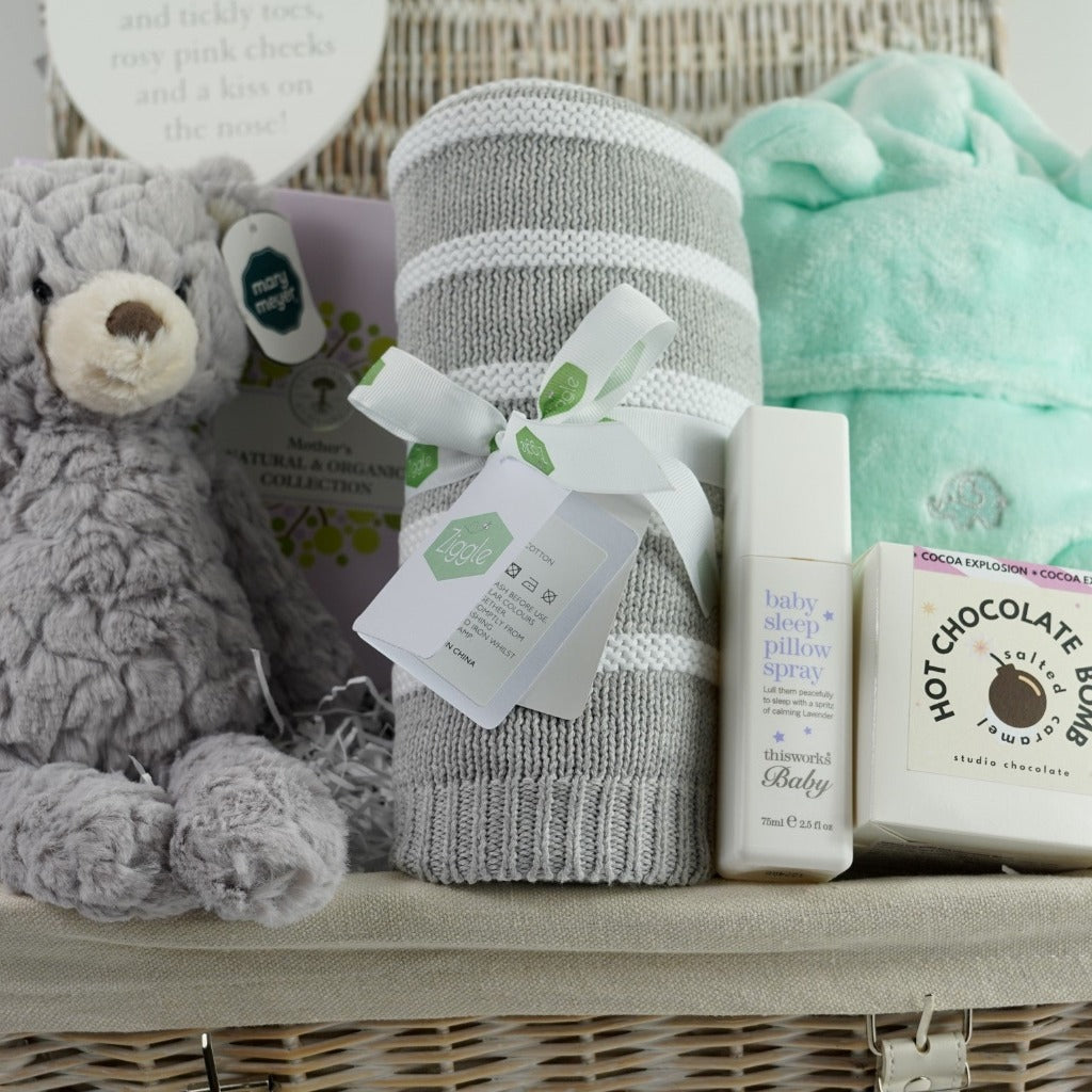 white wicker hamper basket, Neals ayard Organic Mum toiletries, Grey soft baby teddy, mint green baby dressing gown with cute ears, hot chocolate bomb, baby sleep pillow spray, grey and white stripe ziggle baby blanket