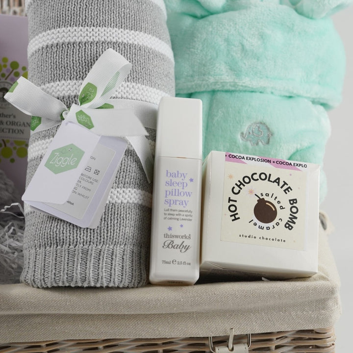 white wicker hamper basket, Neals ayard Organic Mum toiletries, Grey soft baby teddy, mint green baby dressing gown with cute ears, hot chocolate bomb, baby sleep pillow spray, grey and white stripe ziggle baby blanket