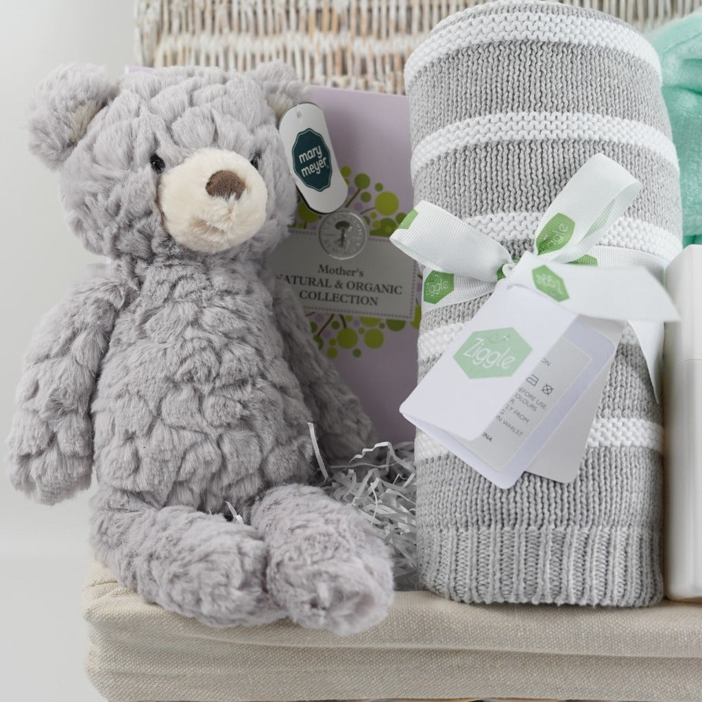 white wicker hamper basket, Neals ayard Organic Mum toiletries, Grey soft baby teddy, mint green baby dressing gown with cute ears, hot chocolate bomb, baby sleep pillow spray, grey and white stripe ziggle baby blanket