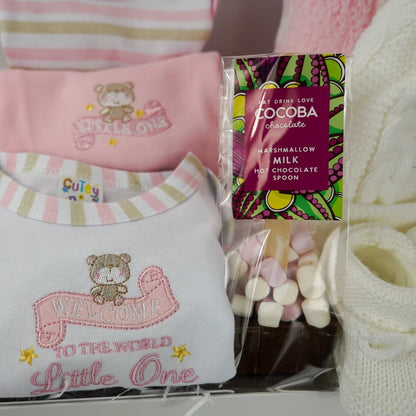 baby girl hamper box with 3 baby vesys with cute designs and pink trim, hot chocolate spoon with marshmallows, baby white cable pom pom hat, white knit booties, pink knit booties , pink baby blanket with white teddy faces