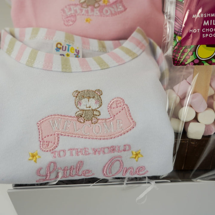 baby girl hamper box with 3 baby vesys with cute designs and pink trim, hot chocolate spoon with marshmallows, baby white cable pom pom hat, white knit booties, pink knit booties , pink baby blanket with white teddy faces