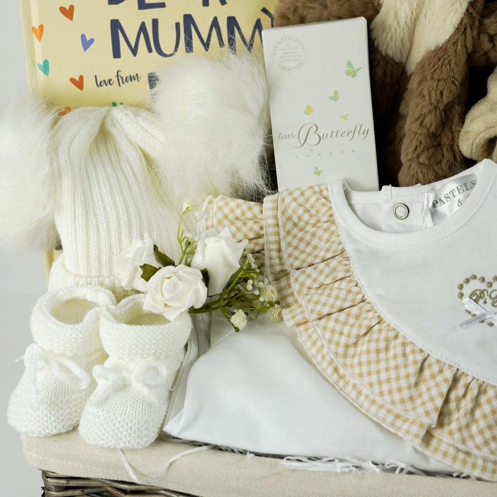Natural hamer basket with baby gifts includes luxury baby clothing in white with caramel check ruffles, baby book, soft teddy comforter, baby dressing gown with ears, fluffy hat with double pom poms in white, natural baby toiletries, knitted booties , baby natural rubber bath toy/teether in Girafee shape