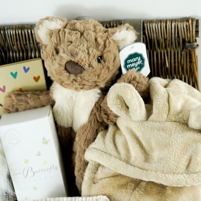 Antique washed hamper basket with Portuguese baby sleepsuit, giraffe rubber teether and bath toy in yellow, white knit bootees, white double fluffy pom pom hat, Dear mummy book, teddy comforter soft toy, butterscotch coloured dressing gown with ears