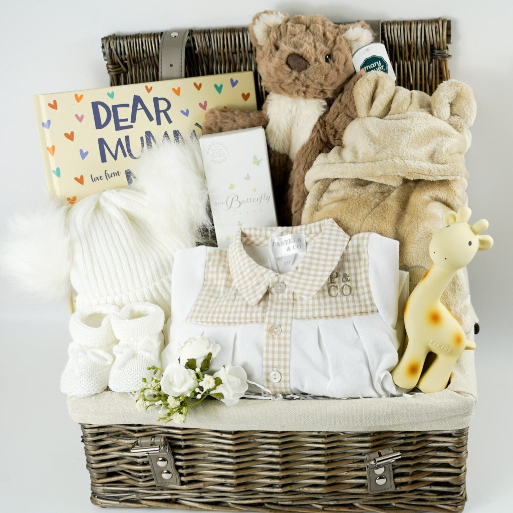 Antique washed hamper basket with Portuguese baby sleepsuit, giraffe rubber teether and bath toy in yellow, white knit bootees, white double fluffy pom pom hat, Dear mummy book, teddy comforter soft toy, butterscotch coloured dressing gown with ears  