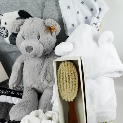White wash hamper basket containg new baby gifts including a grey top with a dog face in black and white, black and white baby leggings and matching socks, Grey steiff teddy, natural hairbrush, white baby dressing gown with ears, white baby slippers