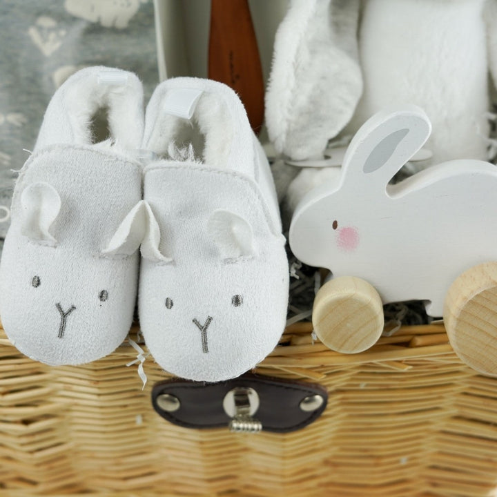 Wicker baby hamper with bunny romper in grey with a white shirt, designer baby hairbrush, baby natural toiletries, wooden rabbit toy on wheels, white baby slippers with a cute face, wooden plaque