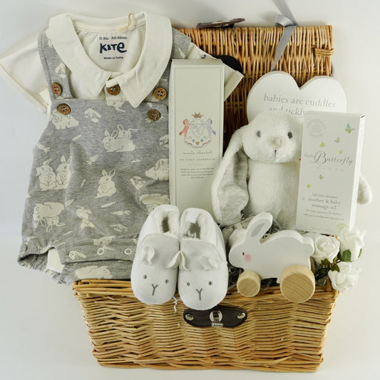 Wicker baby hamper with bunny romper in grey with a white shirt, designer baby hairbrush, baby natural toiletries, wooden  rabbit toy on wheels, white baby slippers with a cute face, wooden plaque 