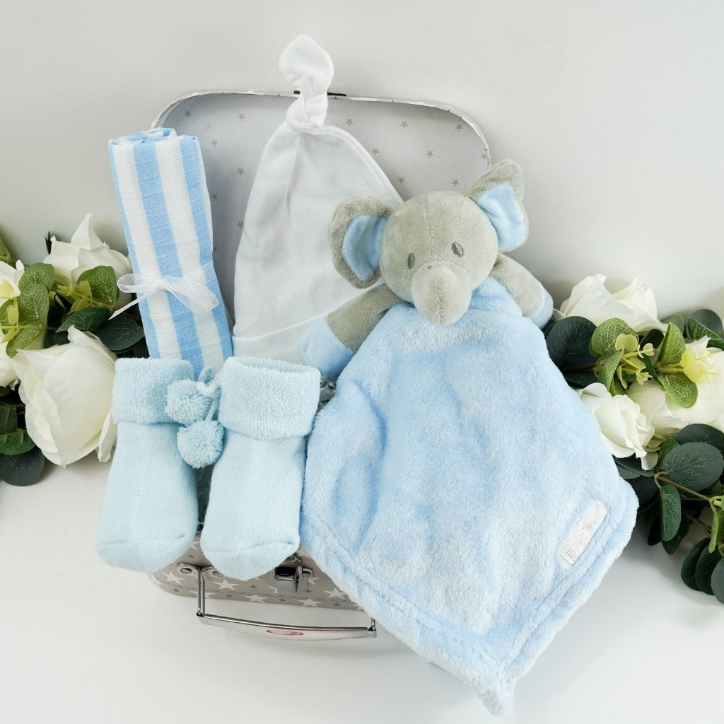 small baby hamper in a grey and white gift case, blue muslin, blue socks with pom poms and cute writing on the sole, baby knot hat in white , soft blue and grey baby elephant comforter 