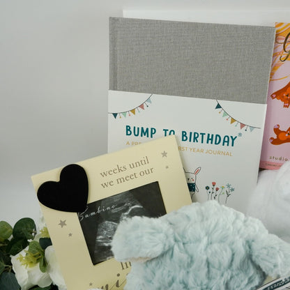 antenatal pregnancy hamper with baby scan photo frame , pale blue/green soft baby teddy, pregnancy and first year of life journal in a grey cover, cute white baby dressing gown with cute ears, white knit baby booties, fairy tale inspired chocolate