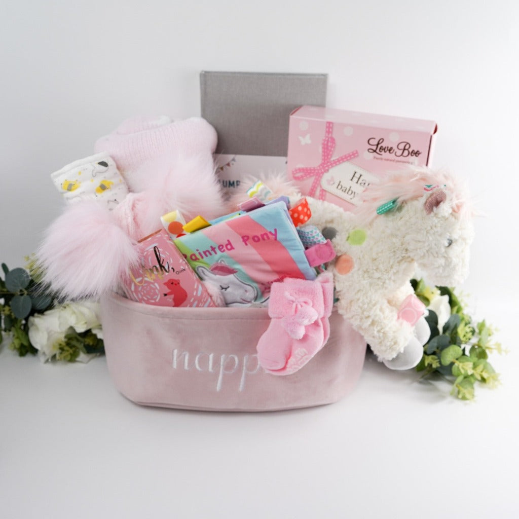 Nappy Caddy in pink full of baby and mum to be gifts, soft pink double pom pom baby hat, baby muslin with elephants, soft pink chenille baby blanket with white heats, pregnancy to first birthday journal, mum and baby natural toiletries , soft white pony with pastel taggies, baby taggie book to match the pony with crinkle effect, studio chocolate bar in gorgeous pink wrapper with gold writing and picture of pigs, pink baby socks 