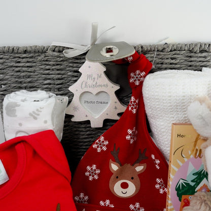 Grey baby hamper with a soft cuddly baby reindeer toy by Mary Meyer, red baby bodysuit with a reindeer and first christmas writing, red knot hat with reindeer and snowklakes and baby first christmas , studio chocolate gingerbread and toffee luxury chocolate bar in luxury wrapping, white knit booties with a tie, white pom pom baby socks , soft muslin in white with winter animals, soft white cellular baby blanket all presented in a grey hamper basket