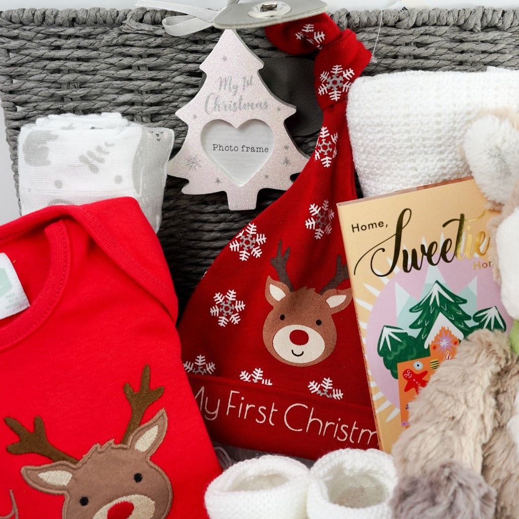 Grey baby hamper with a soft cuddly baby reindeer toy by Mary Meyer, red baby bodysuit with a reindeer and first christmas writing, red knot hat with reindeer and snowklakes and baby first christmas , studio chocolate gingerbread and toffee luxury chocolate bar in luxury wrapping, white knit booties with a tie, white pom pom baby socks , soft muslin in white with winter animals, soft white cellular baby blanket all presented in a grey hamper basket