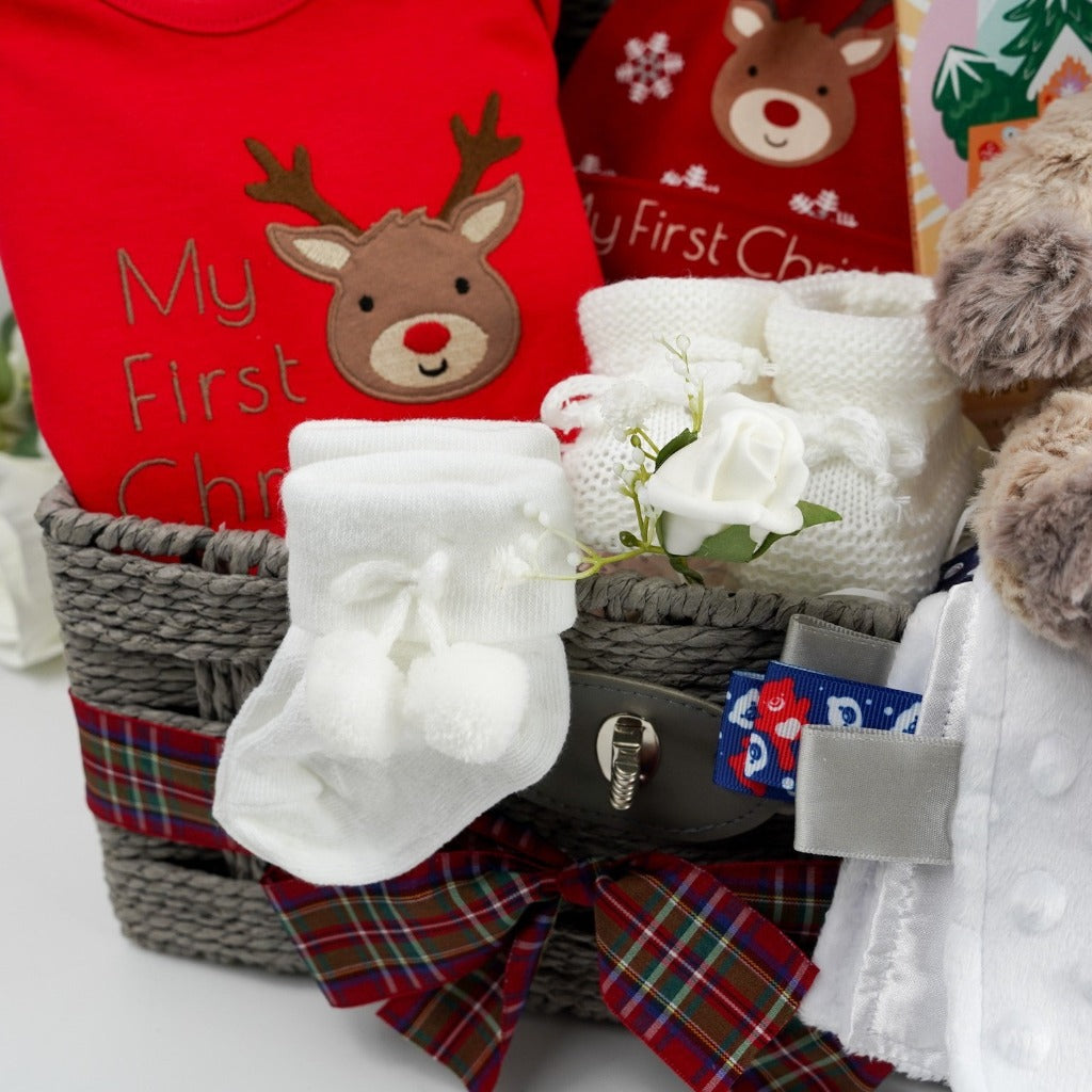 Grey baby hamper with a soft cuddly baby reindeer toy by Mary Meyer, red baby bodysuit with a reindeer and first christmas writing, red knot hat with reindeer and snowklakes and baby first christmas , studio chocolate gingerbread and toffee luxury chocolate bar in luxury wrapping, white knit booties with a tie, white pom pom baby socks , soft muslin in white with winter animals, soft white cellular baby blanket all presented in a grey hamper basket