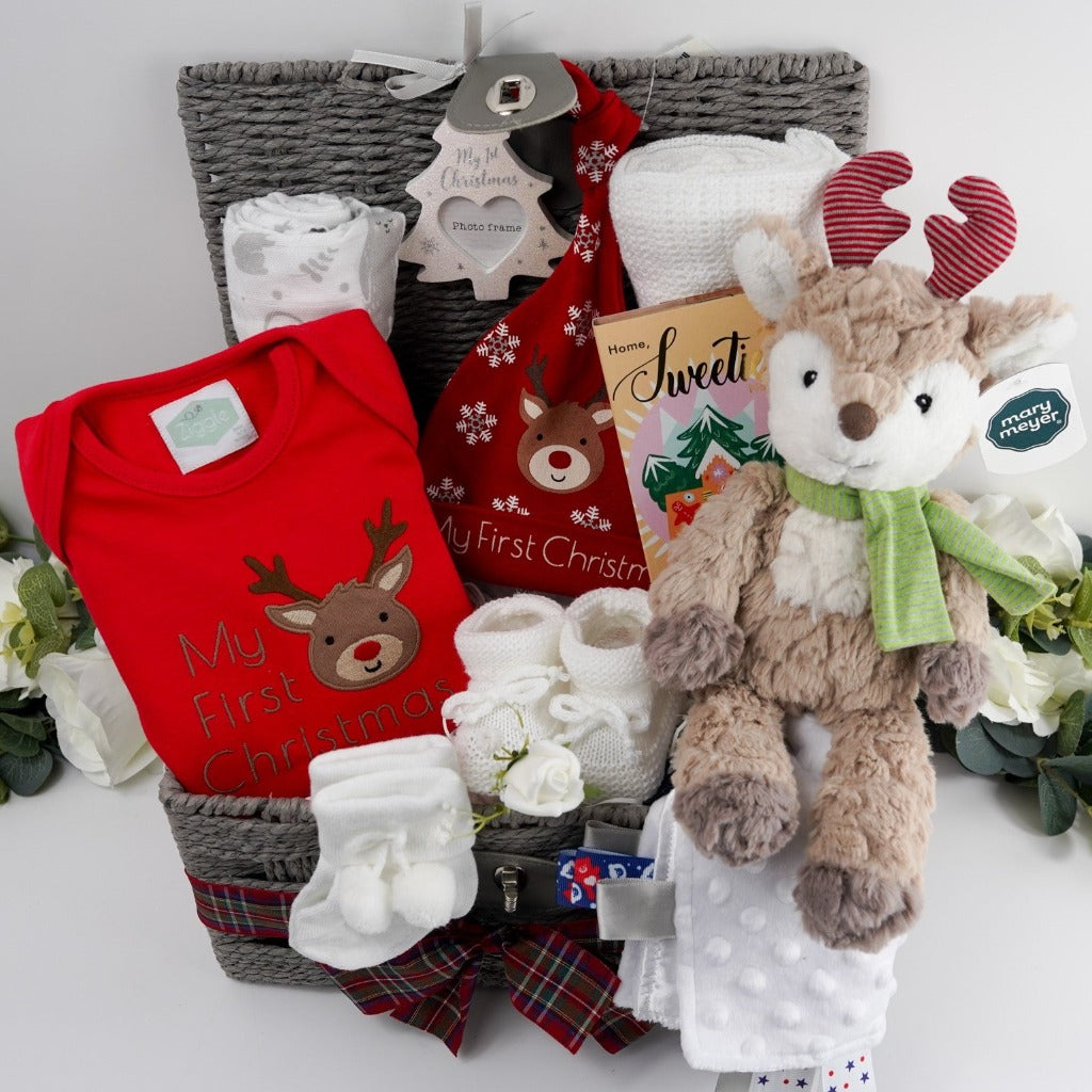 Grey baby hamper with a soft cuddly baby reindeer toy by Mary Meyer, red baby bodysuit with a reindeer and first christmas writing, red knot hat with reindeer and snowklakes and baby first christmas , studio chocolate gingerbread and toffee luxury chocolate bar in luxury wrapping, white knit booties with a tie, white pom pom baby socks , soft muslin in white with winter animals, soft white cellular baby blanket all presented in a grey hamper basket