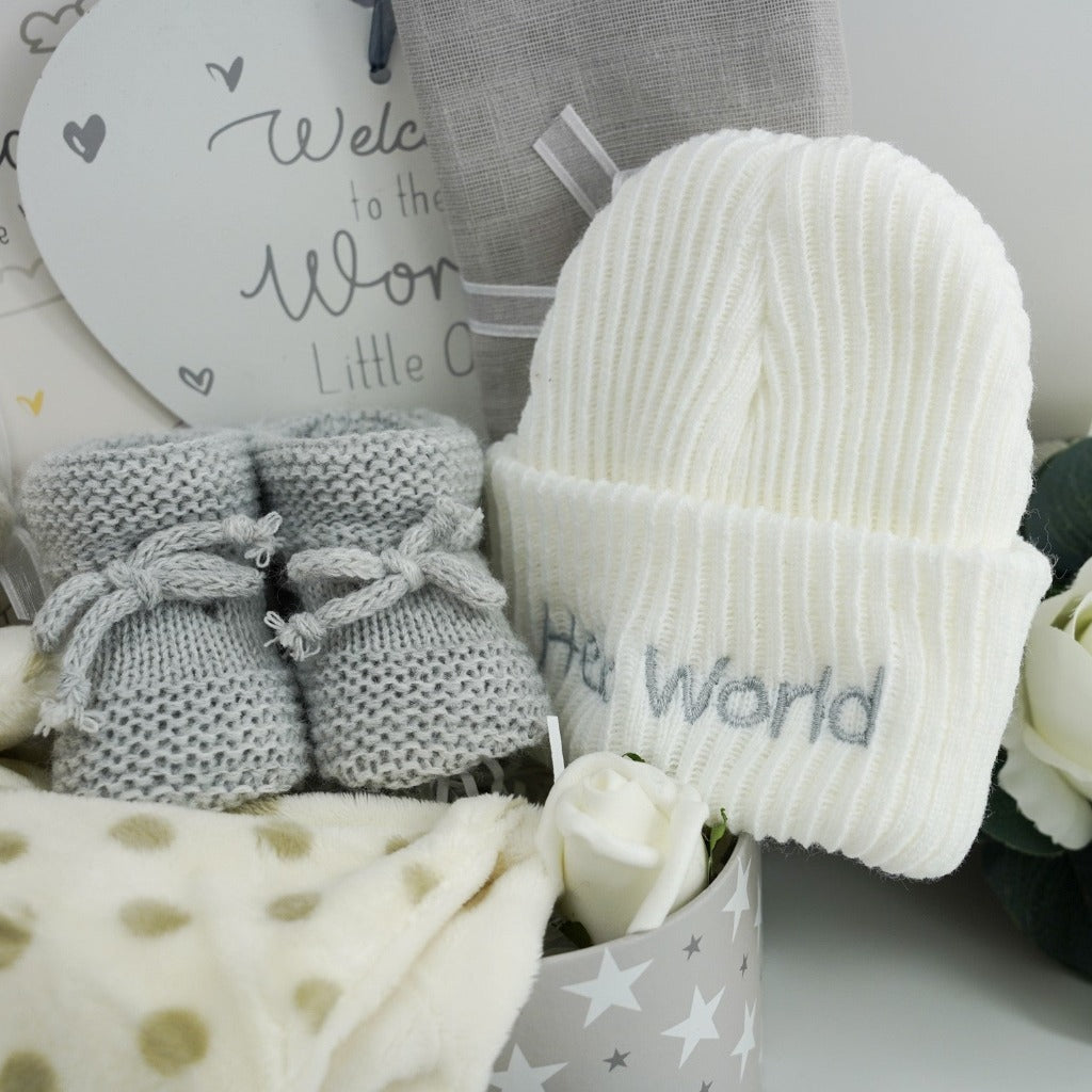 Cream spot giraffe comforter in a neutral colour with a rattle inside, white baby beanie ribbed hat with welcome to the world written on, grey knitted booties with a tie, memorable moments cards , white plaque for the nursery with silver writing welcome to the world , soft grey muslin
