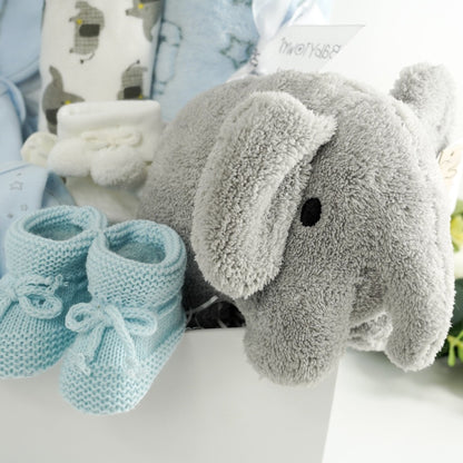 Elephant themed baby gift including a blue 100% cotton baby set with grey elephants, white hooded baby towel with grey elephants, soft pale blue baby blanket with elephants and stars , white baby pom pom socks, soft blue kitted baby bootied , grey terry miffy elephant soft baby toy all in a white hamper box