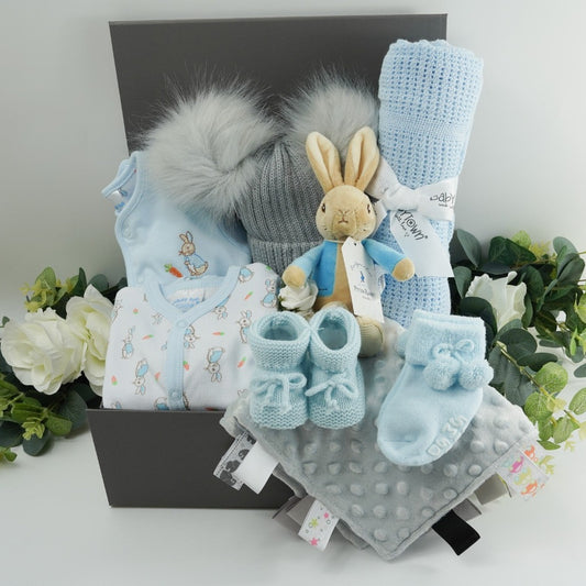Grey ribbon tabbed hamper box with baby gifts, Peter  rabbit clothing set including a Peter Rabbit white sleepsuit with peter rabbit design and pale blue trim, pale blue jacket with Peter rabbit embroidered, soft grey double pom pom baby hat, p[ale blue cellular baby blanket, soft Peter Rabbit first toy with a rattle, pale blue tie up knitted booties, pale blue double pom pom hat, grey taggie comforter 