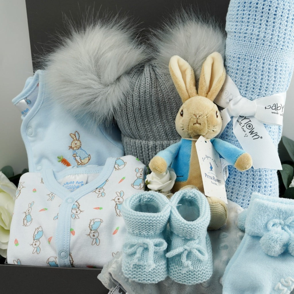 Grey ribbon tabbed hamper box with baby gifts, Peter rabbit clothing set including a Peter Rabbit white sleepsuit with peter rabbit design and pale blue trim, pale blue jacket with Peter rabbit embroidered, soft grey double pom pom baby hat, p[ale blue cellular baby blanket, soft Peter Rabbit first toy with a rattle, pale blue tie up knitted booties, pale blue double pom pom hat, grey taggie comforter