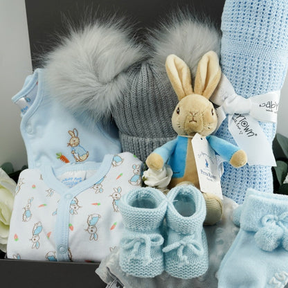 Grey ribbon tabbed hamper box with baby gifts, Peter rabbit clothing set including a Peter Rabbit white sleepsuit with peter rabbit design and pale blue trim, pale blue jacket with Peter rabbit embroidered, soft grey double pom pom baby hat, p[ale blue cellular baby blanket, soft Peter Rabbit first toy with a rattle, pale blue tie up knitted booties, pale blue double pom pom hat, grey taggie comforter