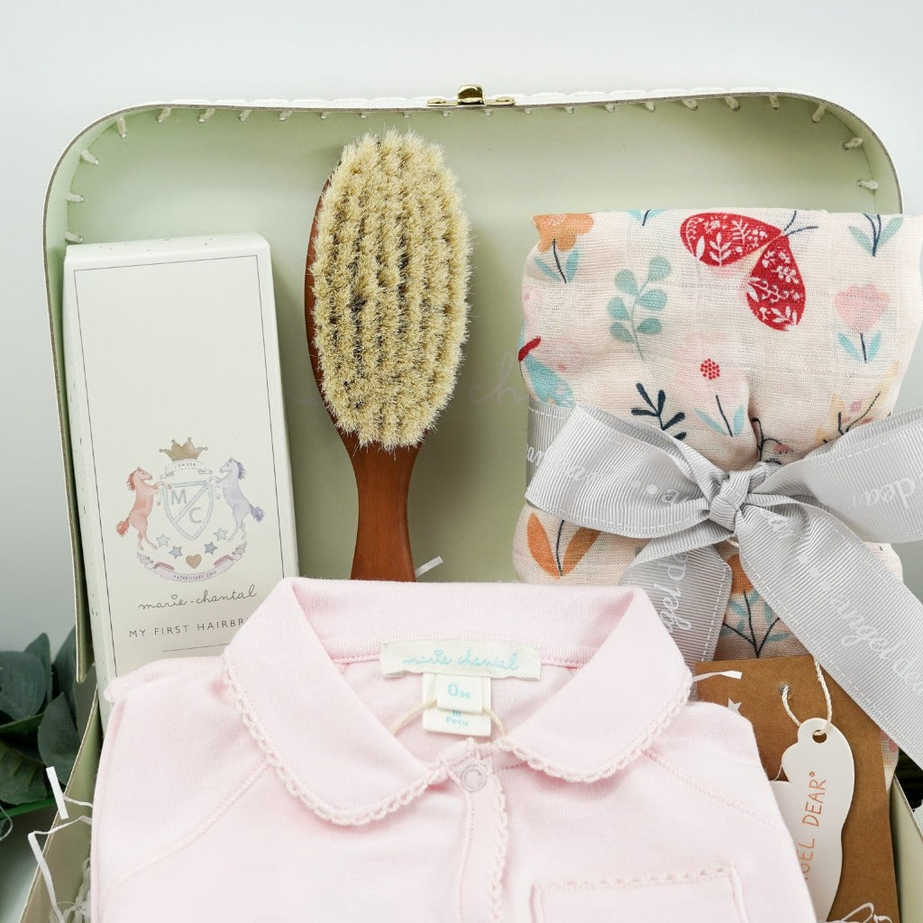 Pale green baby gift suitcase by Marie- Chantal, pink prmium cottom baby sleepsuit with angel wings embroidered on the pocket and picot edging, natural wooden baby hair brush, baby swaddle muslin in pink with butterfly design tied with a ribbon