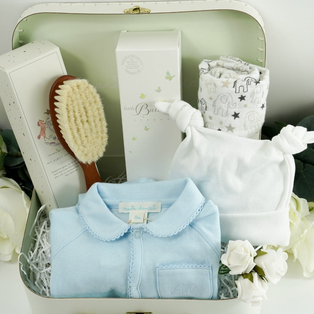 baby storage case with luxury designer baby sleepsuit in pale blue with a picot edge, baby double knot white cotton hat , ziggle muslin in white with grey and black elephants and stars, butterly London baby toiletries in a white box with butterfly design, Marie Chantal baby soft bristle hairbrush in keepsake box