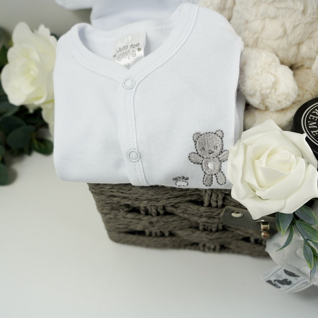 Neutral baby hamper gift in a grey paper rope hamper basket, white cotton baby set includes a white baby sleepsuit with a silver grey teddy embroidered, matching white cotton jacket / cardigan with smocking and grey teddies embroidered, white know hat with silver stars, soft cream plush teddy suitable for a new baby, soft grey bubble baby comforter with ribbon tabs, white knit booties with a tie, grey baby blanket with grey and white teddy heads design, Neal's Yard baby barrier cream in a jarl