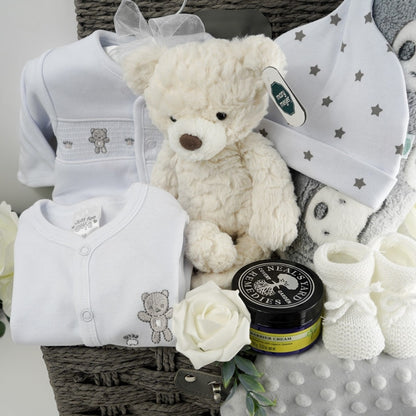 Neutral baby hamper gift in a grey paper rope hamper basket, white cotton baby set includes a white baby sleepsuit with a silver grey teddy embroidered, matching white cotton jacket / cardigan with smocking and grey teddies embroidered, white know hat with silver stars, soft cream plush teddy suitable for a new baby, soft grey bubble baby comforter with ribbon tabs, white knit booties with a tie, grey baby blanket with grey and white teddy heads design, Neal's Yard baby barrier cream in a jarl