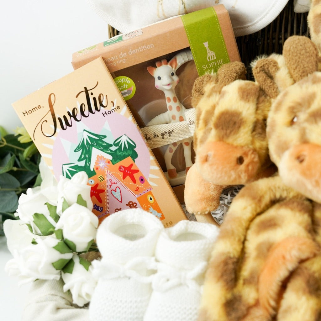 Luxury wicker hamper basket filled with baby gifts including baby clothing set in cream with a giraffe , includes baby sleepsuit, baby vest, baby bib, baby mittens, baby hat, Sophie La Giraffe So Pure rubber teething toy boxed, soft cream baby blanket with giraffe, giraffe soft toy, giraffe comforter, giraffe rattle ring toy, luxury gingerbread toffe chocolate in luxury wrapper, baby knit booties in white with tie, memorable moments baby cards with pastel animals