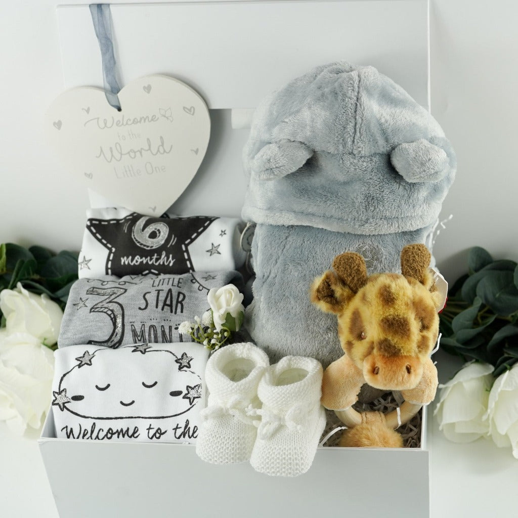 white baby hamper in a ribbon tabbed magnetic box, includes a soft grey baby dressing gown with cute ears baby three milestone vests in white, grey and black , marking baby's arrib=vasl, 3 months and 6 months, white knit booties, giraffe soft rattle and white nursery plaque with welcome to the world in silver writing