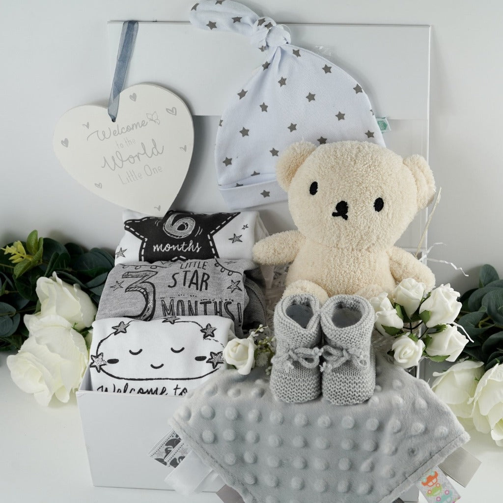 White magnetic tabbed hamper box with baby items including 3 milestone vests, whoite knot baby hat with grey stars , welcome to the world baby plaqye in cream and silver, silver grey baby comforter with ribbon tabs, soft grey knit baby booties , cream terry boris bear from the Miffy range 