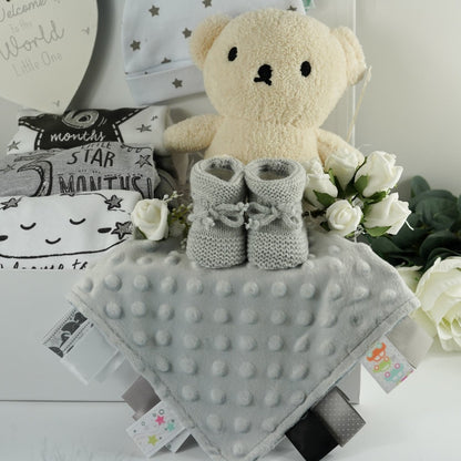 White magnetic tabbed hamper box with baby items including 3 milestone vests, whoite knot baby hat with grey stars , welcome to the world baby plaqye in cream and silver, silver grey baby comforter with ribbon tabs, soft grey knit baby booties , cream terry boris bear from the Miffy range