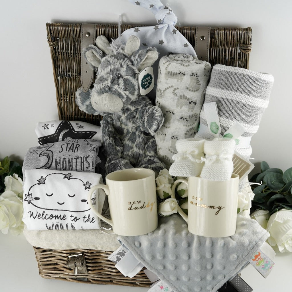 Natural wicker hamper with white lining filled with new parent and baby gifts including, a new mum bone china mug in cream with gold writing, a new  daddy mug in cream with gold writing, 3 milestone baby vests, soft grey giraffe lovie, half toy half comforter, large baby swaddle muslin in white with grey cats, grey and white stripe ziggle heavy baby blanket, soft white knit booties with ties, ribbon tabbed baby comforter in grey 