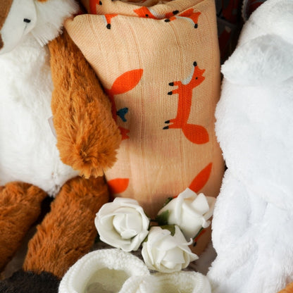 Natural hamper basket with baby items including a fox soft toy with a bushy tale, matching fox soft comforter/finger puttet , large luxery fox muslin in orage with foxes, grey knot hat with orange foxes, grey bandana bub with orange foxes, soft whit knit booties with ties, white velour fleece baby dressing gown with cute ears
