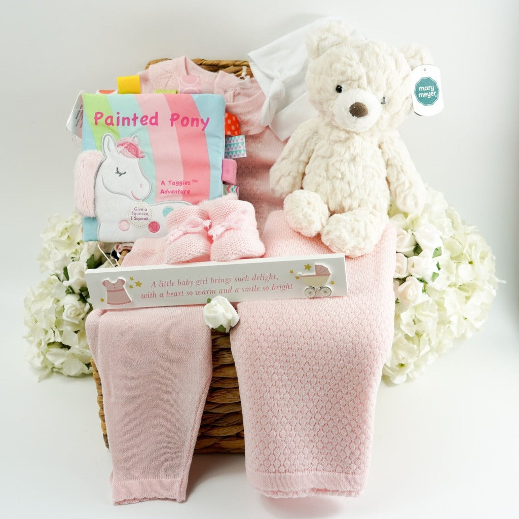 Pink knitted baby set, including pink baby fine knit jumper with frilled collar, knitted baby bottoms in pale pink, matching pale pink shawl, pink knitted baby booties, cream soft teddy bear, painted pony colourful taggie soft book, in hyacinth basket 