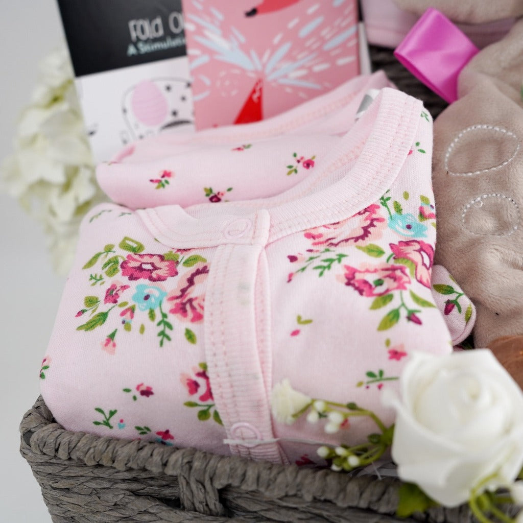 grey hamper basket with a soft fawn lovey with taggies, pink stripe baby blanket heavy weight, baby balck and white book, pink knitted booties, floral baby set, studio chocolate bar
