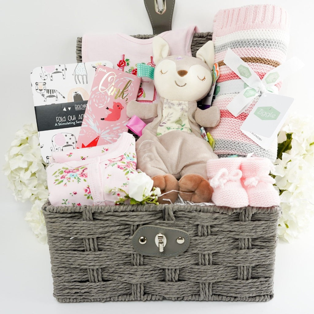 grey hamper basket with a soft fawn lovey with taggies, pink stripe baby blanket heavy weight, baby balck and white book, pink knitted booties, floral baby set, studio chocolate bar