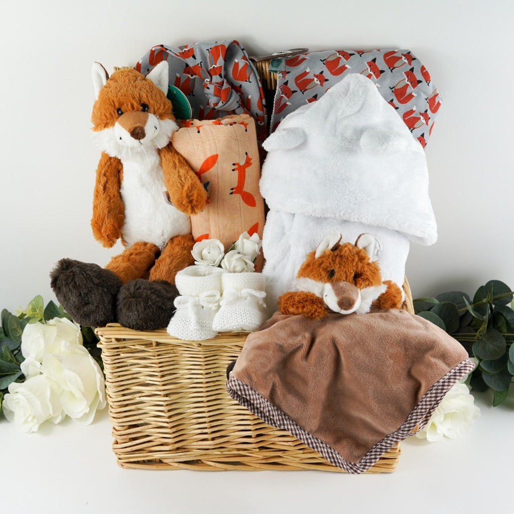 Natural hamper basket with baby items including a fox soft toy with a bushy tale, matching fox soft comforter/finger puttet , large luxery fox muslin in orage with foxes, grey knot hat with orange foxes, grey bandana bub with orange foxes, soft whit knit booties with ties, white velour fleece baby dressing gown with cute ears