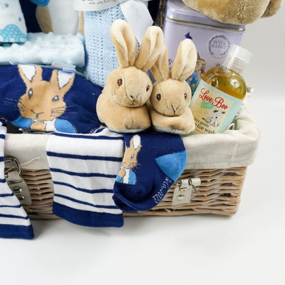 Peter rabbit themed baby basket with navy clothing set with a top with PPeter Rabbit applique design, stiped leggings with Peter Rabbit on the bottom, matching softs, Peter Rabbit soft slippers, Muslin in white with green and blue dinosaurs, pale blue cotton cellular blanket, soft Peter Rabbit toy, Tin of tea with Peter Rabbit design, Natural baby toiletries