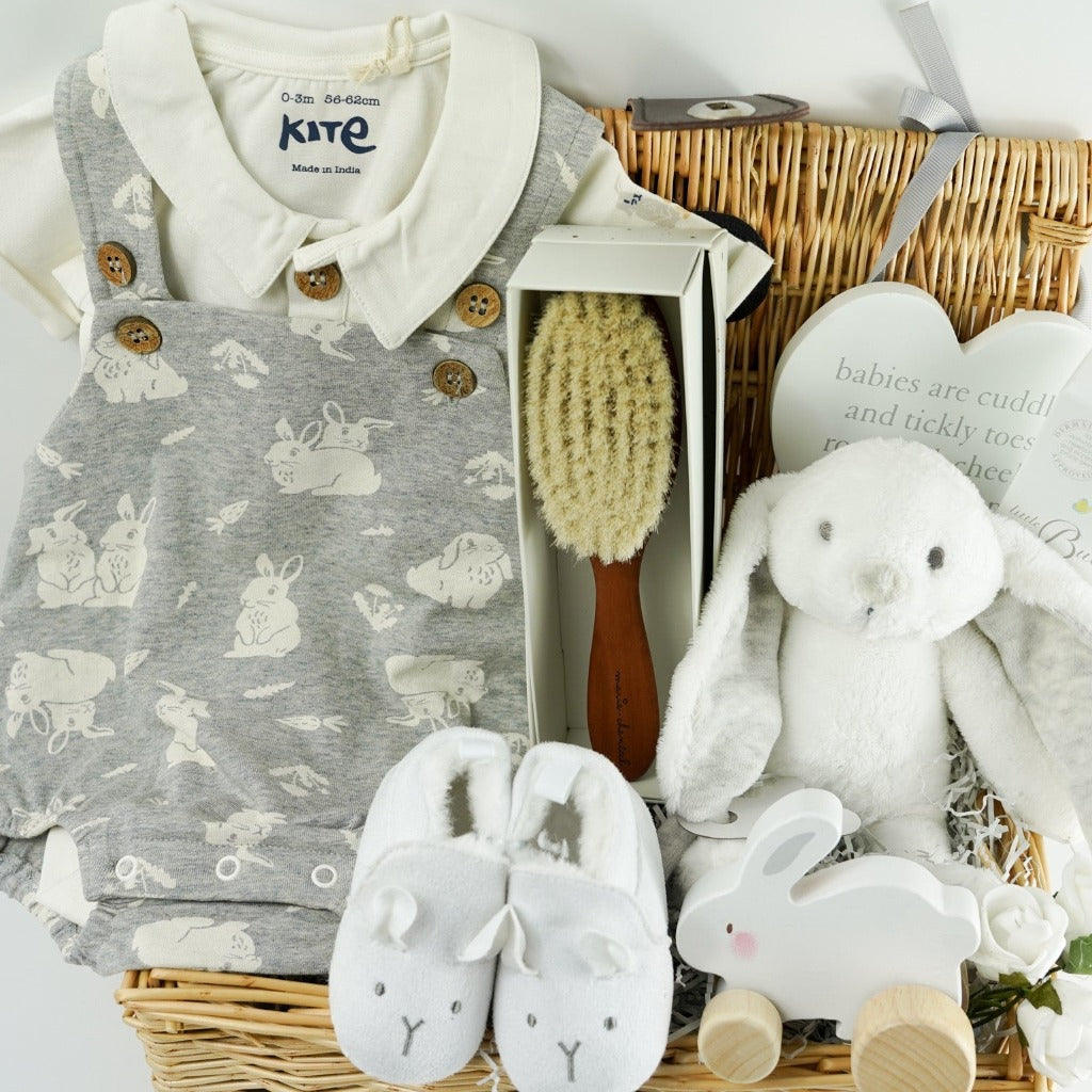 Wicker baby hamper with bunny romper in grey with a white shirt, designer baby hairbrush, baby natural toiletries, wooden rabbit toy on wheels, white baby slippers with a cute face, wooden plaque