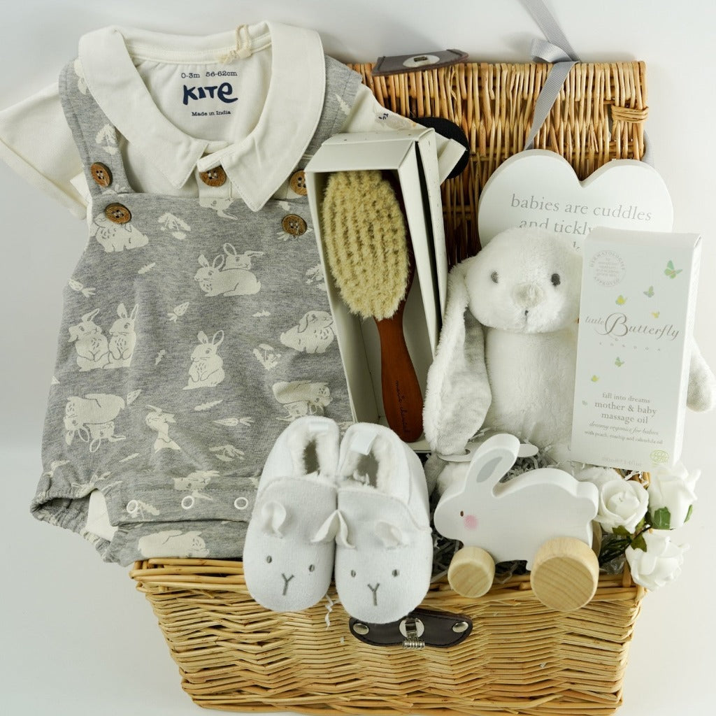 Wicker baby hamper with bunny romper in grey with a white shirt, designer baby hairbrush, baby natural toiletries, wooden rabbit toy on wheels, white baby slippers with a cute face, wooden plaque