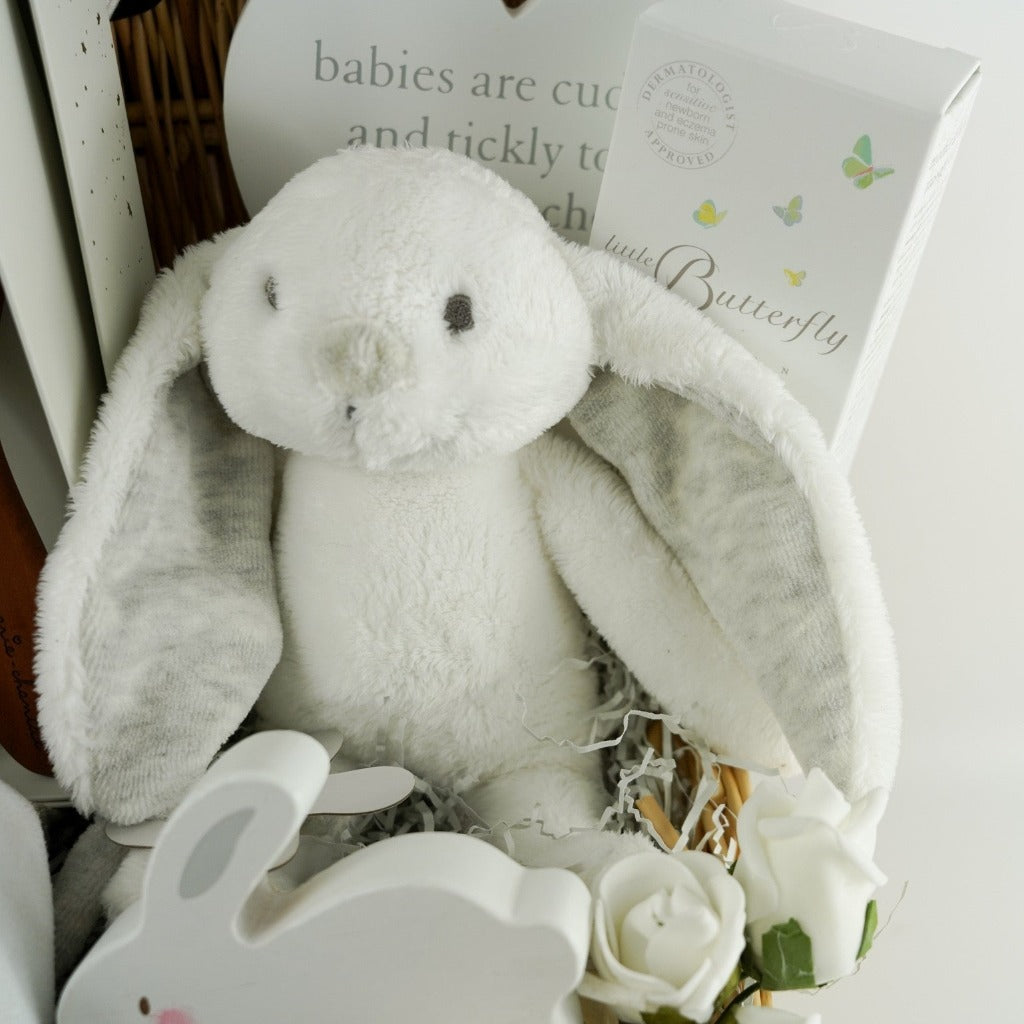 Wicker baby hamper with bunny romper in grey with a white shirt, designer baby hairbrush, baby natural toiletries, wooden rabbit toy on wheels, white baby slippers with a cute face, wooden plaque